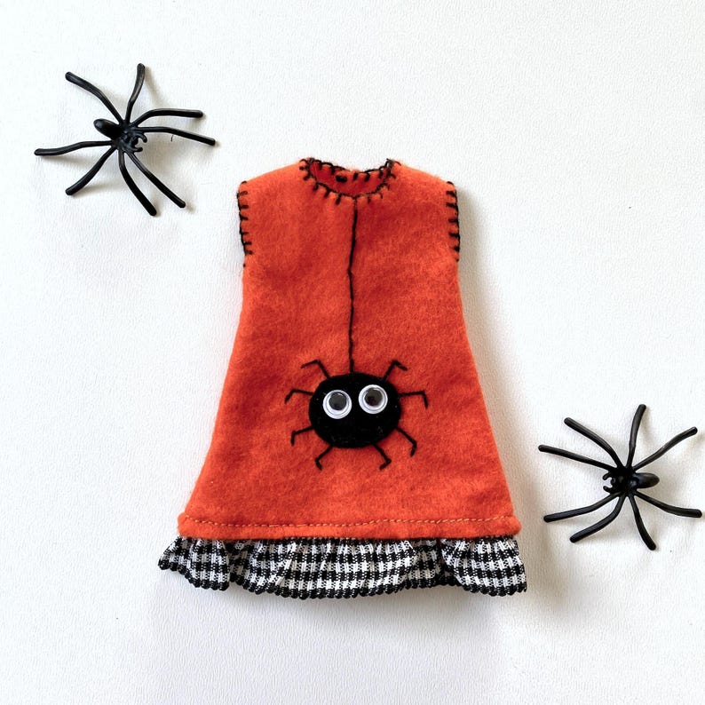 Puede incluir: Un vestido de fieltro naranja con un dise&ntilde;o de ara&ntilde;a negra y un dobladillo a cuadros blanco y negro. Dos ara&ntilde;as de pl&aacute;stico negro se colocan a cada lado del vestido. El vestido tiene costuras negras.