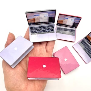 May include: A collection of miniature laptops in shades of silver, red, and pink. Each laptop has a screen showing a desktop interface and a keyboard. The laptops are held in a hand, highlighting their small size and intricate design.