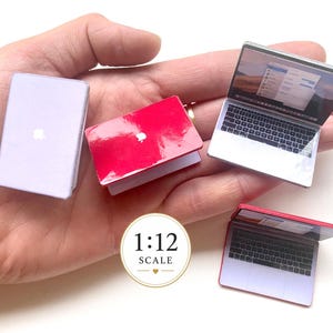 Könnte beinhalten: Eine Sammlung von Miniatur-Laptop-Modellen im Maßstab 1:12, bestehend aus einem geschlossenen hellvioletten Laptop, einem geschlossenen roten Laptop und zwei geöffneten Laptops mit sichtbaren Bildschirmen und Tastaturen. Jedes Gerät hat ein kleines weißes Logo.