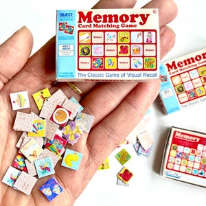 Peut inclure: Un jeu de mémoire miniature avec une boîte rouge, blanche et bleue. La boîte porte l'inscription "Memory Card Matching Game". Le jeu comprend 32 cartes avec différentes images. Les cartes sont éparpillées sur une main.