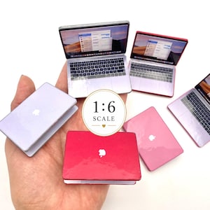 Puede incluir: Una colección de modelos de portátiles en miniatura a escala 1:6 en varios colores, incluyendo plata, rojo y rosa. Cada portátil presenta un pequeño logotipo blanco en la tapa y una pantalla abierta que muestra una interfaz digital.