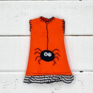 Puede incluir: Decoraci&oacute;n de Halloween de fieltro naranja con forma de vestido, con una ara&ntilde;a negra con ojos de bot&oacute;n. El vestido tiene un volante a cuadros blanco y negro en la parte inferior y costuras negras.