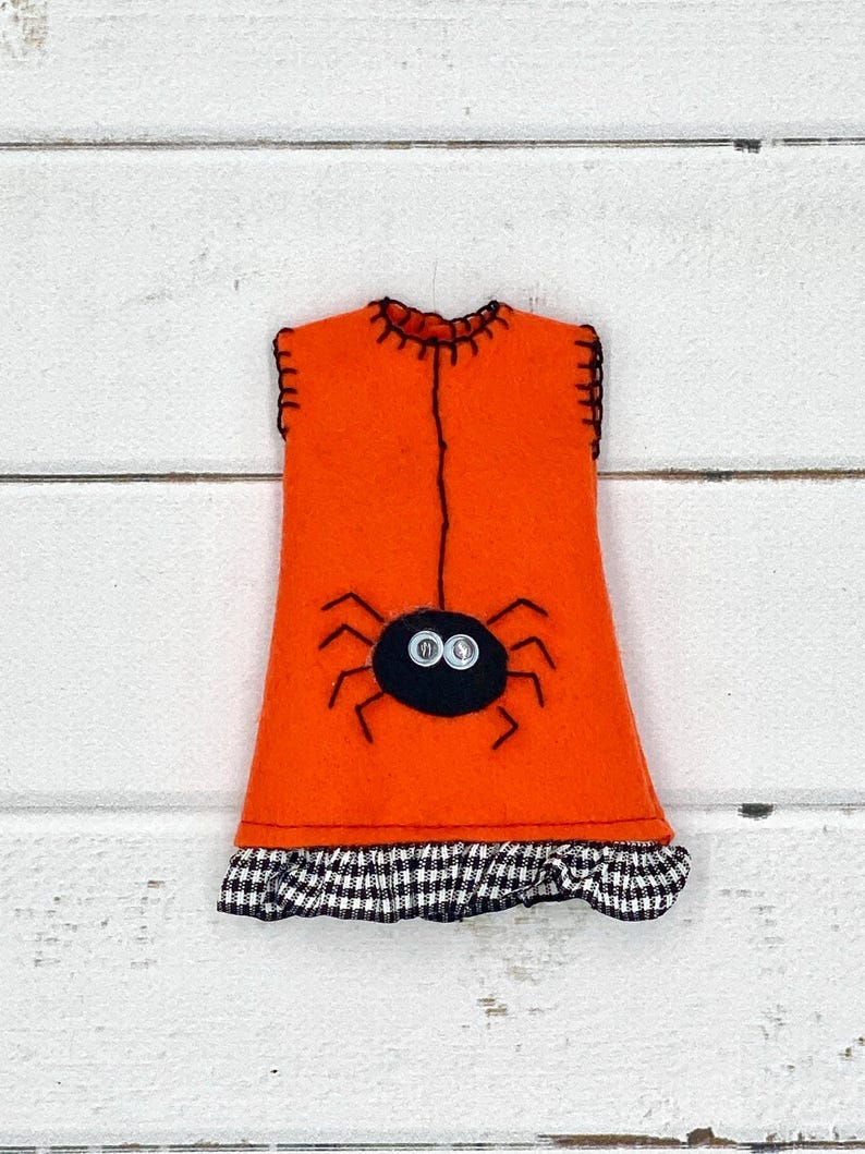 Puede incluir: Decoraci&oacute;n de Halloween de fieltro naranja con forma de vestido, con una ara&ntilde;a negra con ojos de bot&oacute;n. El vestido tiene un volante a cuadros blanco y negro en la parte inferior y costuras negras.