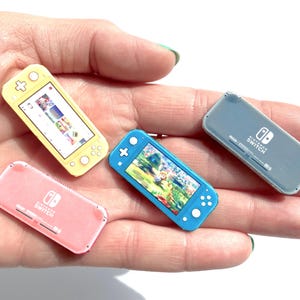 Peut inclure: Miniatures de consoles Nintendo Switch jaunes, bleues, roses et grises. Les consoles jaunes et bleues affichent des écrans de jeu. Les consoles roses et grises affichent le logo Nintendo Switch. Ces petits appareils de jeu sont tenus dans une main.
