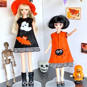 Puede incluir: Dos mu&ntilde;ecas vestidas con disfraces de Halloween. Una mu&ntilde;eca lleva un vestido negro con un fantasma y un murci&eacute;lago, un sombrero de bruja naranja y botas negras. La otra mu&ntilde;eca lleva un vestido naranja con una ara&ntilde;a y zapatos negros. Decoraciones de Halloween en el fondo.