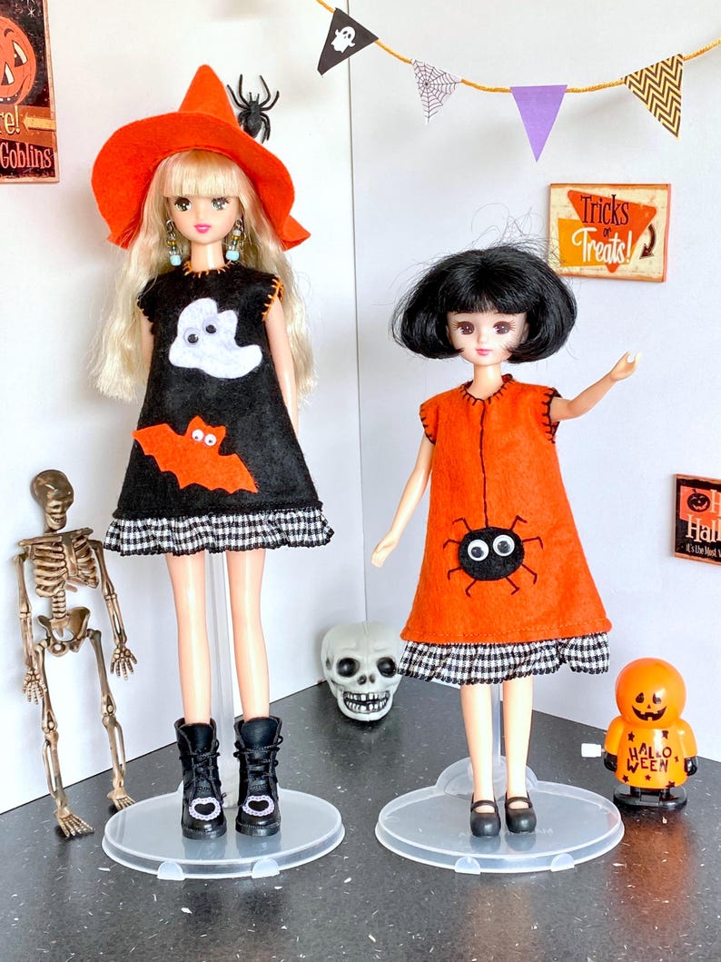 Puede incluir: Dos mu&ntilde;ecas vestidas con disfraces de Halloween. Una mu&ntilde;eca lleva un vestido negro con un fantasma y un murci&eacute;lago, un sombrero de bruja naranja y botas negras. La otra mu&ntilde;eca lleva un vestido naranja con una ara&ntilde;a y zapatos negros. Decoraciones de Halloween en el fondo.