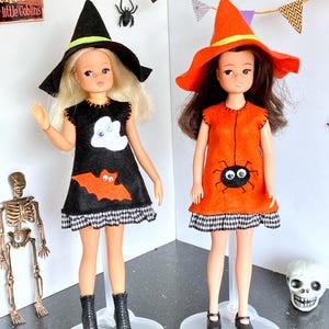 Puede incluir: Dos mu&ntilde;ecas vestidas para Halloween. Una mu&ntilde;eca lleva un vestido negro con un fantasma y un murci&eacute;lago, un sombrero de bruja negro y botas negras. La otra mu&ntilde;eca lleva un vestido naranja con una ara&ntilde;a, un sombrero de bruja naranja y zapatos negros. Ambas mu&ntilde;ecas est&aacute;n sobre bases transparentes.