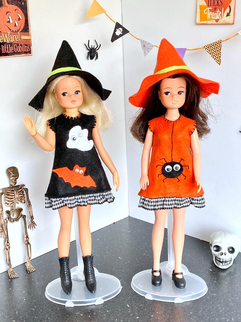 Puede incluir: Dos mu&ntilde;ecas vestidas para Halloween. Una mu&ntilde;eca lleva un vestido negro con un fantasma y un murci&eacute;lago, un sombrero de bruja negro y botas negras. La otra mu&ntilde;eca lleva un vestido naranja con una ara&ntilde;a, un sombrero de bruja naranja y zapatos negros. Ambas mu&ntilde;ecas est&aacute;n sobre bases transparentes.