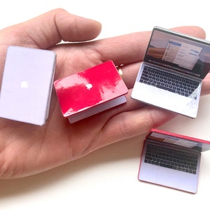 May include: Four miniature laptop computers, two with silver cases and two with red cases. The laptops are arranged on a hand.