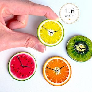 Könnte beinhalten: Vier runde Miniaturuhren mit Fruchtscheiben-Designs, darunter Limette, Kiwi, Wassermelone und Orange. Jedes Zifferblatt zeigt die Zahlen eins bis zwölf und schwarze Zeiger. Eine kreisförmige Grafik oben rechts zeigt die Aufschrift 1:6 SCALE.