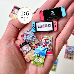 Peut inclure: Une console de jeu vidéo miniature à l'échelle 1:6 avec un écran noir et des manettes bleue et rose, accompagnée de plusieurs boîtiers de jeu miniatures présentant des illustrations pour des titres comme Zelda, Mario Odyssey et Pokemon Legends Arceus.