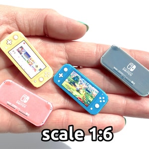 Puede incluir: Cuatro consolas Nintendo Switch en miniatura de diferentes colores: amarillo, rosa, azul y gris. Las consolas se muestran en una mano, con el texto "escala 1:6" debajo.