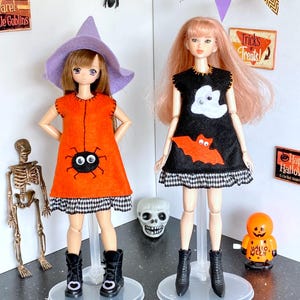 Puede incluir: Dos mu&ntilde;ecas vestidas con disfraces de Halloween. Una mu&ntilde;eca lleva un vestido naranja con un dise&ntilde;o de ara&ntilde;a y un sombrero de bruja morado. La otra mu&ntilde;eca lleva un vestido negro con un dise&ntilde;o de fantasma y murci&eacute;lago. Ambas mu&ntilde;ecas llevan botas negras y est&aacute;n sobre bases transparentes.