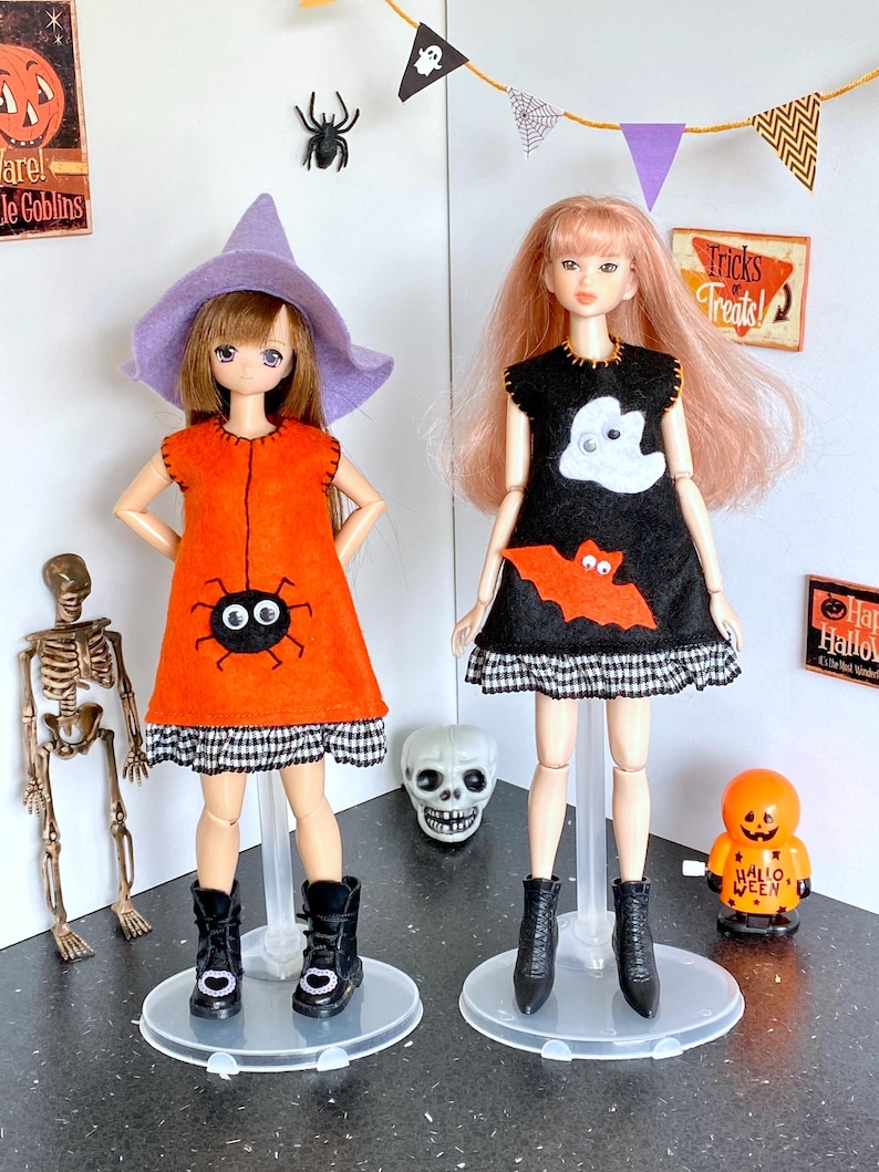 Puede incluir: Dos mu&ntilde;ecas vestidas con disfraces de Halloween. Una mu&ntilde;eca lleva un vestido naranja con un dise&ntilde;o de ara&ntilde;a y un sombrero de bruja morado. La otra mu&ntilde;eca lleva un vestido negro con un dise&ntilde;o de fantasma y murci&eacute;lago. Ambas mu&ntilde;ecas llevan botas negras y est&aacute;n sobre bases transparentes.