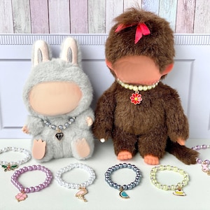 May include: Two plush dolls, a grey bunny and a brown monkey, each with a pearl necklace. Several beaded bracelets with charms are displayed in front of the dolls. The bracelets have charms such as a flamingo, a flower, and a watermelon.