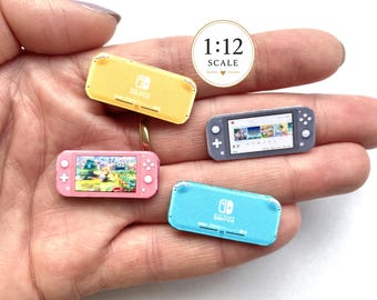 May include: Four miniature handheld game consoles in 1:12 scale. The collection includes a yellow and a light blue console showing the back, a pink console displaying a game screen, and a grey console showing the front screen with game icons.