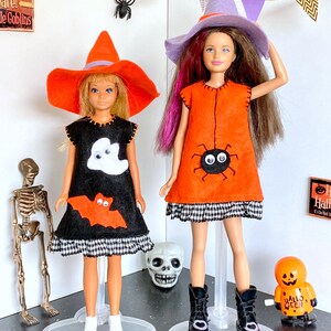 Puede incluir: Dos mu&ntilde;ecas vestidas con trajes de Halloween. Una mu&ntilde;eca lleva un vestido negro con apliques de fantasma y murci&eacute;lago, combinado con un sombrero de bruja naranja. La otra mu&ntilde;eca lleva un vestido naranja con un aplique de ara&ntilde;a y un sombrero de bruja morado.