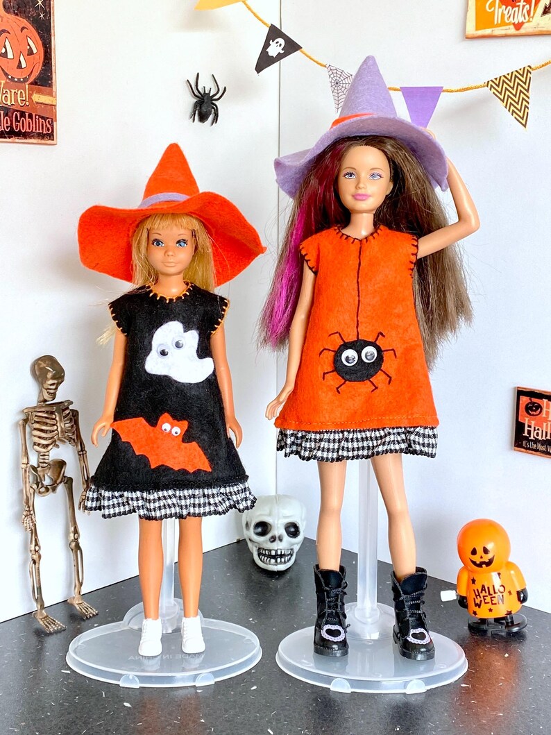 Puede incluir: Dos mu&ntilde;ecas vestidas con trajes de Halloween. Una mu&ntilde;eca lleva un vestido negro con apliques de fantasma y murci&eacute;lago, combinado con un sombrero de bruja naranja. La otra mu&ntilde;eca lleva un vestido naranja con un aplique de ara&ntilde;a y un sombrero de bruja morado.