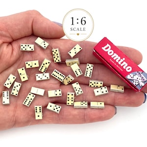 May include: A set of miniature dominoes in a 1:6 scale, featuring off-white rectangular tiles with black pips. The set includes a small red box labelled "Domino Double Six Dominoes" with a graphic of dominoes on the front of the packaging.