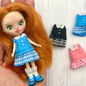 Puede incluir: Una muñeca con un vestido de cuadros azules y blancos con un corpiño azul y un cuello blanco. La muñeca tiene el pelo rojo y los ojos verdes. Se muestran tres vestidos adicionales en el fondo: uno de cuadros azules y blancos, uno de cuadros negros y blancos y uno de cuadros rosas y blancos.