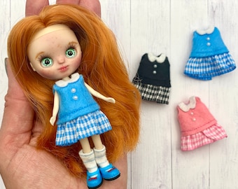 May include: A doll wearing a blue and white gingham dress with a blue bodice and white collar. The doll has red hair and green eyes. Three additional dresses are shown in the background: one blue and white gingham, one black and white gingham, and one pink and white gingham.