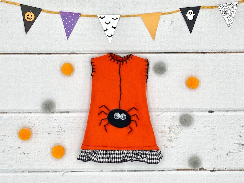 Puede incluir: Vestido de Halloween de fieltro naranja con una aplicaci&oacute;n de ara&ntilde;a negra y un volante a cuadros blanco y negro. El vestido est&aacute; decorado con costuras negras y se exhibe con banderines tem&aacute;ticos de Halloween y pompones naranjas y grises.