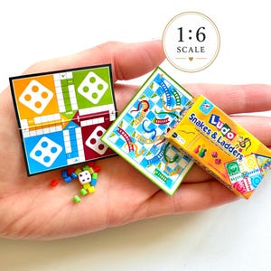 May include: A miniature 1:6 scale board game set featuring a Ludo board with yellow, green, blue, and red quadrants, a Snakes and Ladders board, a small box labeled Ludo Snakes & Ladders, and tiny colored game pieces with a single white die.