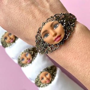 Può includere: Un bracciale a polsino color bronzo con un volto di bambola al centro, con occhi blu e labbra rosa, incastonato in un design a filigrana. Sono visibili ulteriori accenti di volti di bambola.