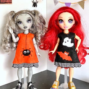 Puede incluir: Dos mu&ntilde;ecas vestidas con trajes de Halloween. Una mu&ntilde;eca lleva un vestido naranja con una ara&ntilde;a, la otra un vestido negro con un fantasma y un murci&eacute;lago. Ambos vestidos tienen un ribete a cuadros blanco y negro. Las mu&ntilde;ecas tienen pelo plateado y rojo, y est&aacute;n sobre soportes transparentes.