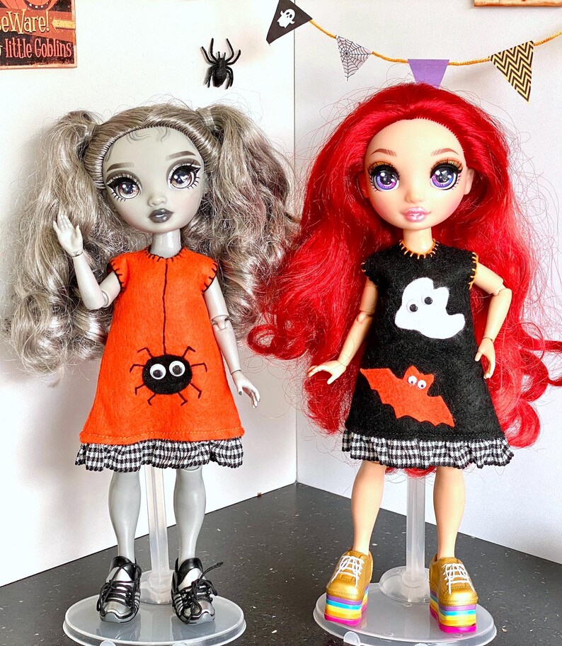 Puede incluir: Dos mu&ntilde;ecas vestidas con trajes de Halloween. Una mu&ntilde;eca lleva un vestido naranja con una ara&ntilde;a, la otra un vestido negro con un fantasma y un murci&eacute;lago. Ambos vestidos tienen un ribete a cuadros blanco y negro. Las mu&ntilde;ecas tienen pelo plateado y rojo, y est&aacute;n sobre soportes transparentes.