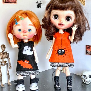 Puede incluir: Dos mu&ntilde;ecas vestidas con trajes de Halloween. Una mu&ntilde;eca lleva un vestido negro con un aplique de fantasma y murci&eacute;lago, zapatillas blancas. La otra mu&ntilde;eca lleva un vestido naranja con un dise&ntilde;o de ara&ntilde;a y botas negras. Decoraciones de Halloween en el fondo.