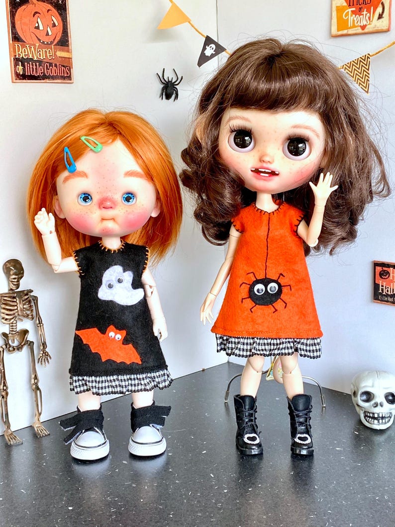 Puede incluir: Dos mu&ntilde;ecas vestidas con trajes de Halloween. Una mu&ntilde;eca lleva un vestido negro con un aplique de fantasma y murci&eacute;lago, zapatillas blancas. La otra mu&ntilde;eca lleva un vestido naranja con un dise&ntilde;o de ara&ntilde;a y botas negras. Decoraciones de Halloween en el fondo.