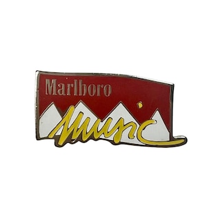 May include: A red and white enamel pin featuring the Marlboro logo and the words "Marlboro Country" in yellow.