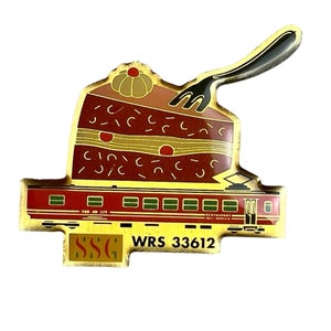 May include: A metal pin featuring a slice of chocolate cake topped with a dollop of cream and a fork, resting above a red train carriage. The train is labelled with the text SSG and WRS 33612, featuring SBB CFF branding and restaurant car markings.
