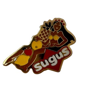May include: A gold-coloured pin featuring a cartoon woman with brown skin, wearing a yellow bikini and a red flower crown, holding a pineapple and other fruit. The pin has the word "Sugus" in white letters on a red background.
