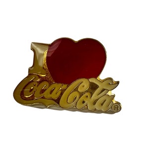 May include: Gold-colored pin with a red heart and the words "I love Coca-Cola" in white lettering with a registered trademark symbol.