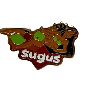 May include: A gold-coloured pin featuring a cartoon image of a woman in a green bikini lying on a red heart-shaped object with the word "Sugus" written in white letters.