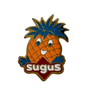 May include: A gold-coloured pin featuring a cartoon pineapple with a smiling face. The pineapple is wearing a blue crown and has a red and white diamond-shaped logo with the word "sugus" in white letters.