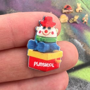 Può includere: Una spilla Playskool con un clown dei cartoni animati con cappello rosso, sciarpa verde e abito blu, seduto su una scatola rossa e gialla. Sullo sfondo sono visibili altre spille con vari disegni.
