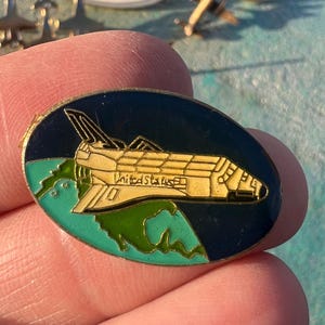 May include: An oval-shaped pin featuring a yellow space shuttle design with the words "United States" on it. The pin has a dark blue background with green accents and is surrounded by other pins with airplane designs.