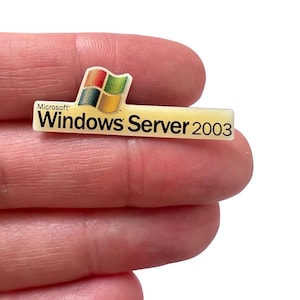May include: A rectangular pin featuring the Microsoft Windows logo and the text "Windows Server 2003". The logo includes a colorful flag design. The pin is cream-colored with black text.