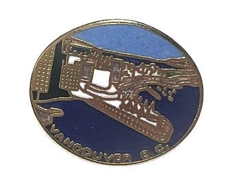 May include: A round metal enamel pin featuring a stylized graphic of the Vancouver, B.C. skyline and harbor. The design includes a light blue sky, dark blue water, and white building silhouettes with the text VANCOUVER B.C. along the bottom edge.