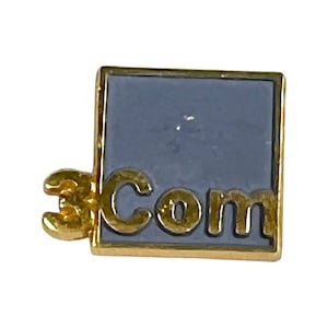 Puede incluir: Un pin de color dorado con fondo azul y el texto "3Com" en letras doradas.