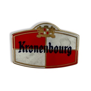 Pode incluir: Um alfinete de metal dourado com um design xadrez vermelho e branco. O alfinete apresenta o texto "Kronenbourg" em letras pretas com contorno azul.