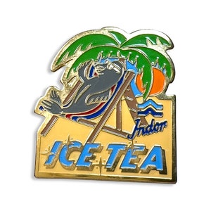 May include: A gold-colored metal pin with a cartoon seal wearing a red and blue swimsuit relaxing on a beach chair under a palm tree. The text "ICE TEA" is on the bottom of the pin.