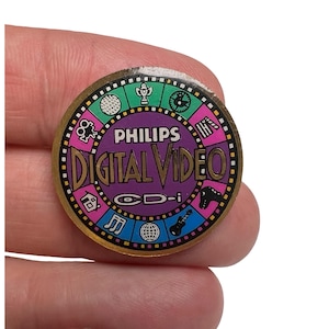 May include: A round, vintage Philips Digital Video CD-i pin. The pin features a gold border with a purple center and various colorful icons. The text "PHILIPS DIGITAL VIDEO" is prominently displayed.