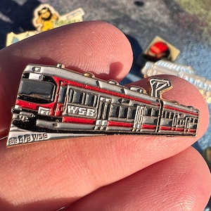 May include: A silver and red enamel pin in the shape of a train. The pin has the letters "WSB" in red on the side. The pin is held between two fingers.