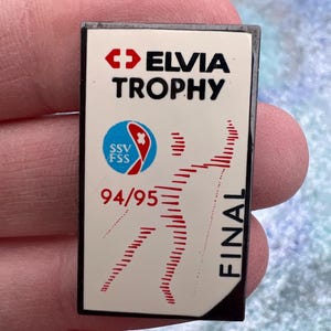 May include: A rectangular pin with the text "ELVIA TROPHY" in black. The pin features a red and white logo, the text "SSV FSS", and the numbers "94/95". Red lines form a stylized figure, with the word "FINAL" in black.