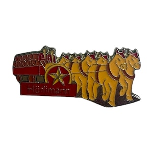 Pode incluir: Um alfinete de metal vermelho e dourado com uma carroça vermelha puxada por seis cavalos dourados com uma estrela na carroça e o texto "Hürlimann" na parte inferior.