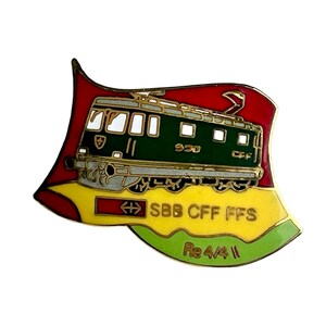 May include: A metal enamel pin shaped like a waving flag featuring a green electric locomotive. The design includes the text SBB CFF FFS and Re 4/4 II in gold lettering against red, yellow, and light green background sections with gold trim.